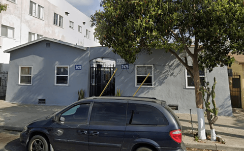 A grey car is parked in front of a grey building with a tree in front of it.