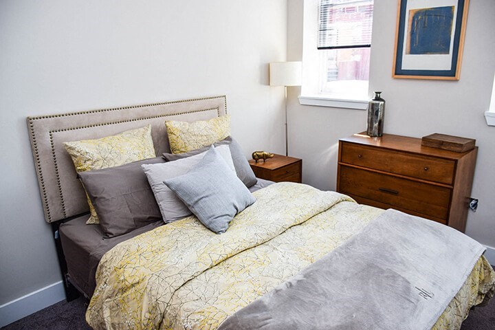 A bed with a yellow comforter and grey pillows sits in a room with a wooden nightstand and a small table beside it.