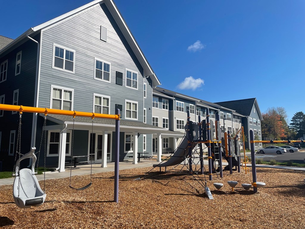 A playground with a swing set and a slide in front of a grey building.