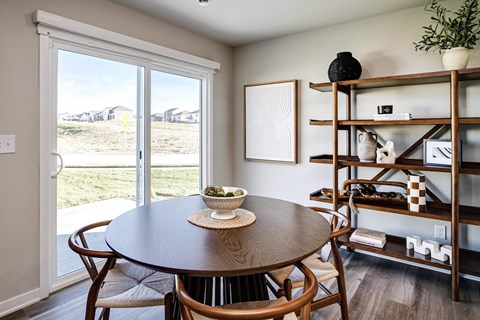 A dining room with a round table and chairs, a shelf with decorative items, and a view of a grassy area outside.