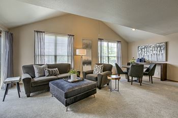 A living room with a grey couch, a grey ottoman, a grey armchair, a grey coffee table, a grey side table, a grey lamp, a grey painting, a grey rug, and a grey carpet.