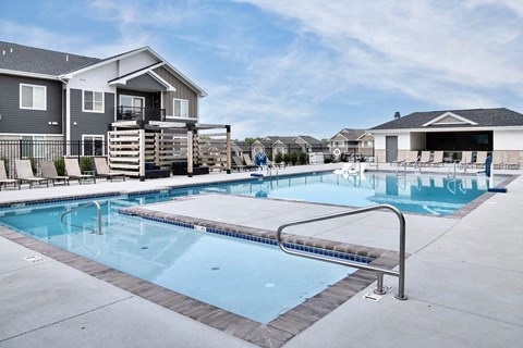 A swimming pool in front of a house with a deck and lounge chairs.