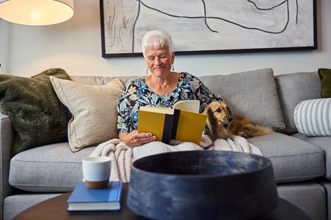 A woman is reading a book while sitting on a couch with a dog beside her.