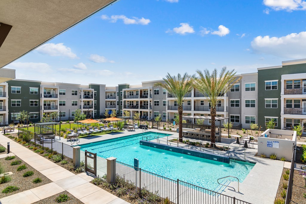 A swimming pool is surrounded by a fence and palm trees in front of apartment buildings.