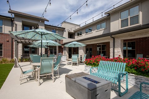 a patio with tables chairs and umbrellas and a fire pit with benches at a senior apartment community