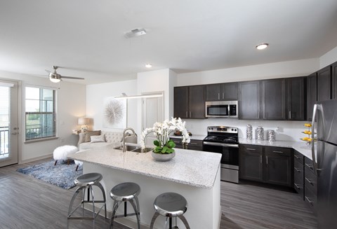a kitchen with stainless steel appliances, granite countertops and island, opening up to a living room at a senior apartment community
