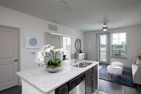 a kitchen with stainless steel appliances, granite countertops and island, opening up to a living room at a senior apartment community