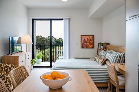 A bowl of oranges sits on a wooden table in a well-lit room with a bed and a television.