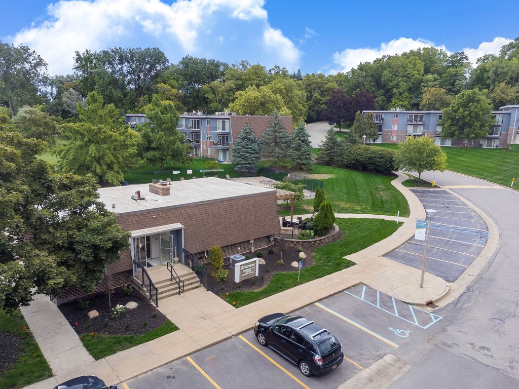 A parking lot with a car parked in the foreground and a building with a covered walkway in the background.