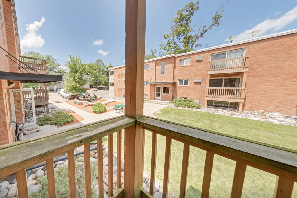 A balcony view of a brick apartment building with a green lawn and trees.