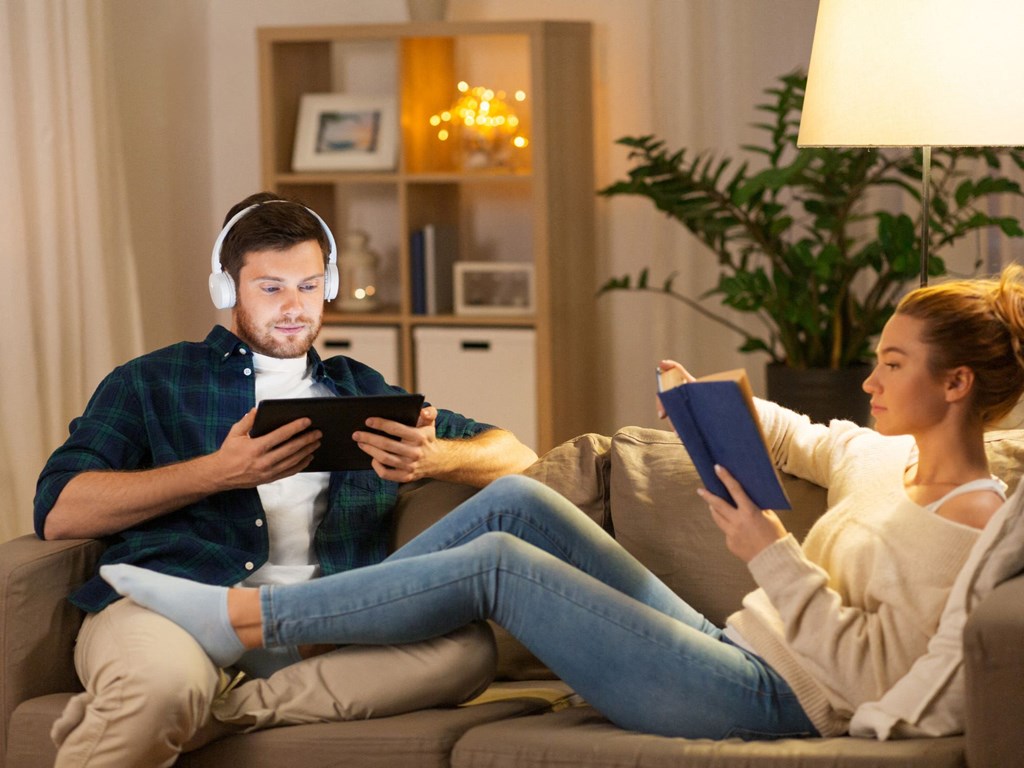 a man and a woman sitting on a couch with headphones on and looking at a tablet