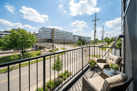 A balcony with a table and chairs overlooks a street with buildings and trees.