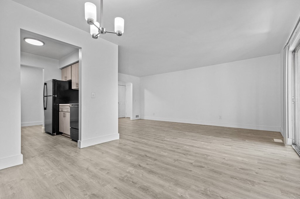 an empty living room with white walls and a kitchen with a black refrigerator