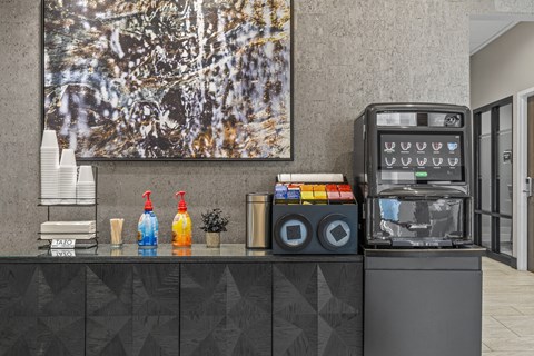 A black and white painting hangs on a wall above a counter with a soda machine and a stack of cups.