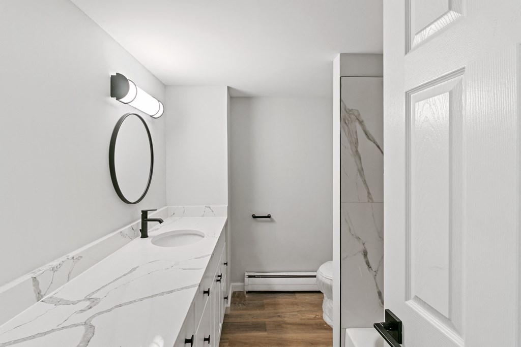 A white bathroom with a marble counter top and a round mirror.