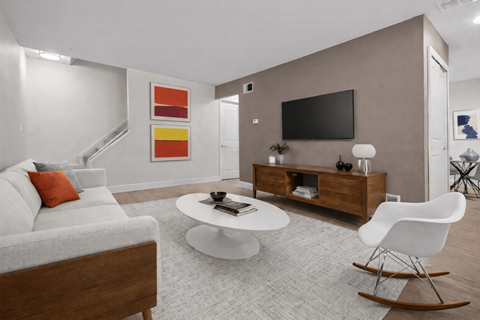 A modern living room with a white sofa, a white coffee table, a white rocking chair, a wooden cabinet, a flat screen TV, and a painting on the wall.
