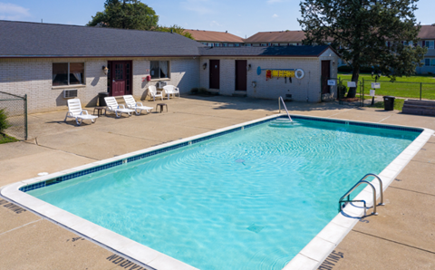 A swimming pool in front of a building with a white fence and lawn chairs.