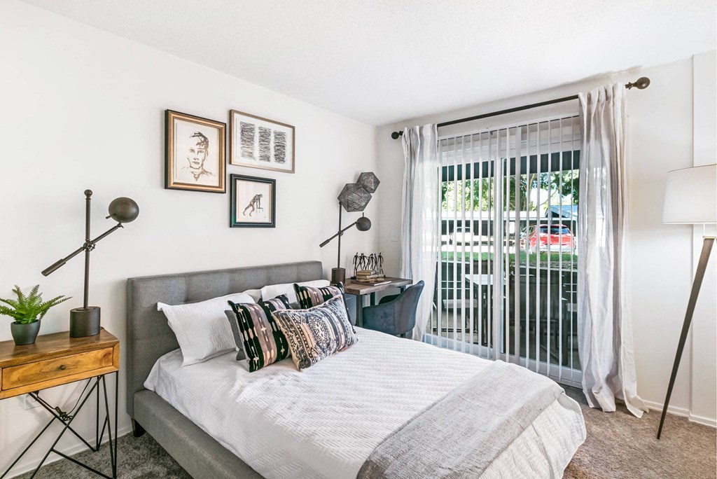 A bedroom with a bed, a nightstand, and a window with a view of a balcony at The Waverly on the Lake - Belleville, MI Apartment , Michigan, 95050