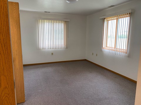 A room with a carpeted floor and two windows with blinds.