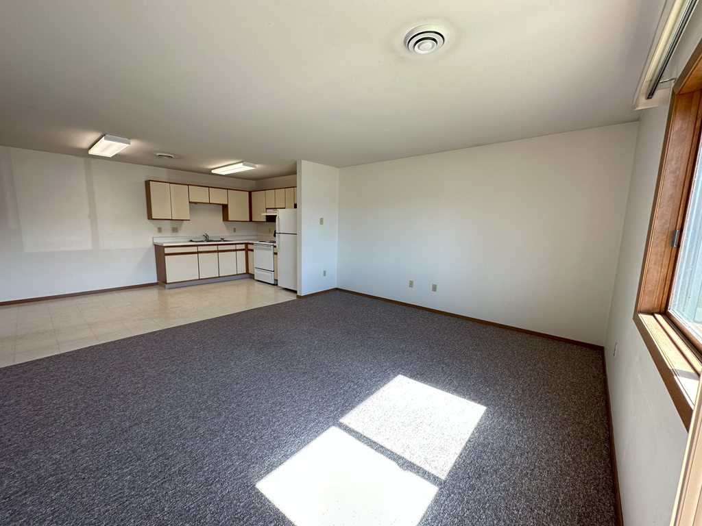 an empty living room with a kitchen in the background