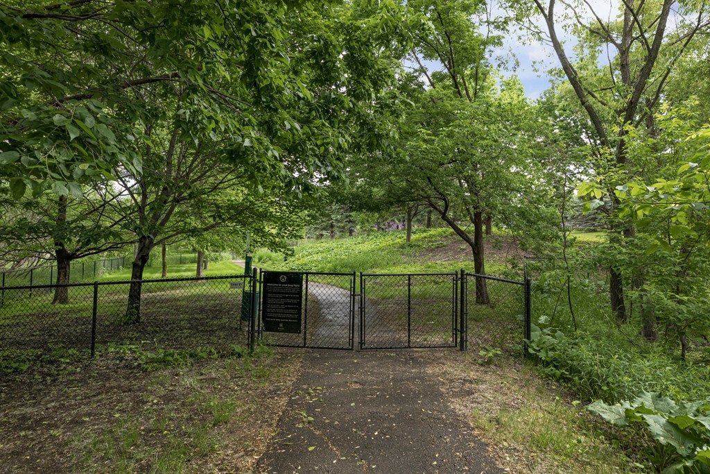 a gate is open to a dog park with trees and a grassy hill in the background  at Shadow Hills, Plymouth, 55442