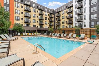 A large swimming pool surrounded by lounge chairs in front of apartment buildings.