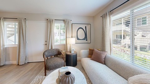 A living room with a grey armchair, a white sofa, a coffee table with a vase on it, and a large window with curtains.