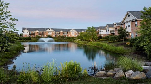 A serene lakeside view with houses in the background and a fountain in the middle.