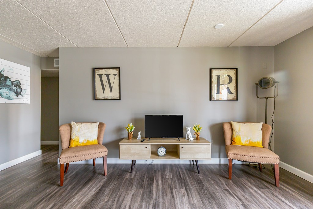 A room with a wooden floor, two chairs, a table with a laptop and a clock.