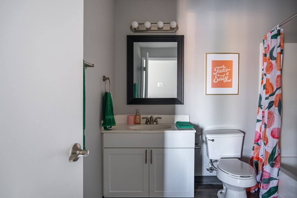 A bathroom with a white toilet, a sink with a mirror above it, and a shower curtain at Centerpointe Apartments, Canandaigua, NY, 14424