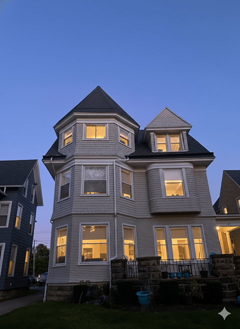 A large house with a tower and multiple windows is lit up at dusk.