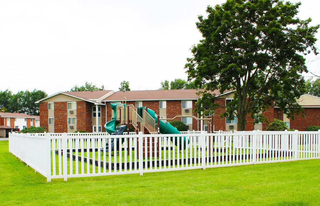 a white fence in front of a brick building with green grass and trees  at Highview Manor Apartments, Fairport,