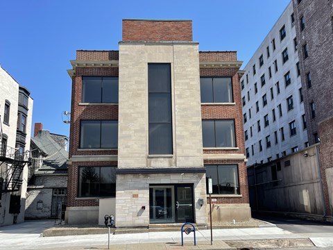 A building with a red brick facade and a large window is surrounded by other buildings.