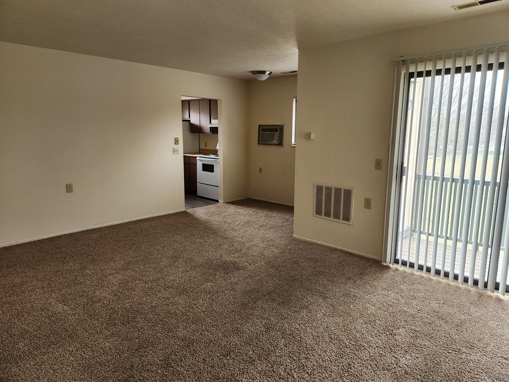 an empty living room with a sliding glass door and a kitchen in the background