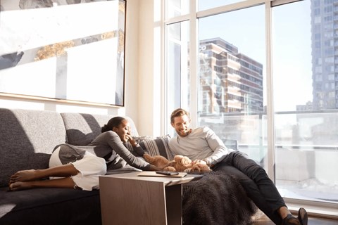 A man and woman are sitting on a couch in a bright room with a large window showing a cityscape.