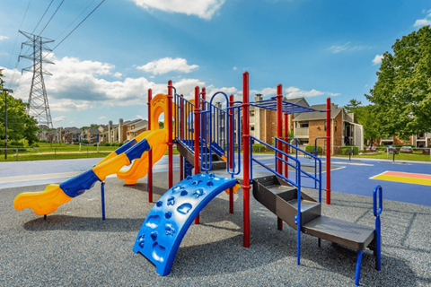 A playground with a blue and yellow slide, red and blue climbing frame, and a grey ramp.