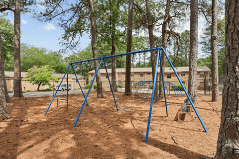 A playground swing set in a wooded area with a building in the background.