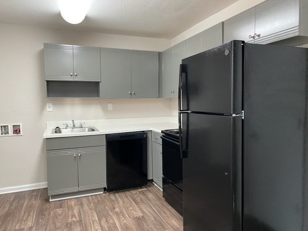 A black refrigerator stands in a kitchen with grey cabinets and a white counter.