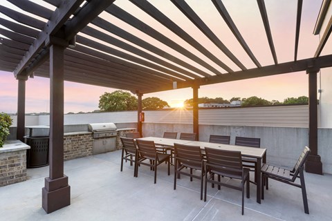 a patio with a wooden table and chairs and a grill with a sunset in the background