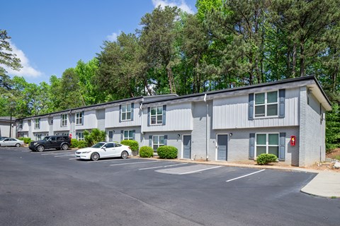 A parking lot in front of a grey building with trees in the background.