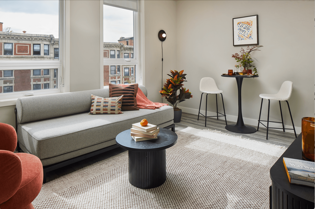A snapshot of the living area in LVL West 1-Bedroom floorplan, featuring a couch against two large windows, a coffee table, and bistro set with two chairs and a table.
