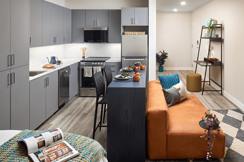A snapshot of a LVL West studio apartment floorplan, looking from the bedroom area into the kitchen and living space. The kitchen features dark gray cabinetry, a while tile backsplash, and light gray flooring. The bar seating leans up against the back of the couch, and there is a small office area with a desk.