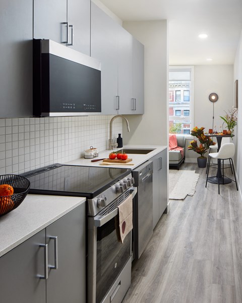 A photo of a galley kitchen looking towards the living room in the LVL West 1-Bedroom floorplan. The kitchen features gray cabinetry, a white backsplash, and light gray laminated flooring.