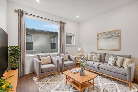 A living room with a grey sofa, a grey armchair, a wooden coffee table, a white rug, a painting on the wall, and a window with curtains.