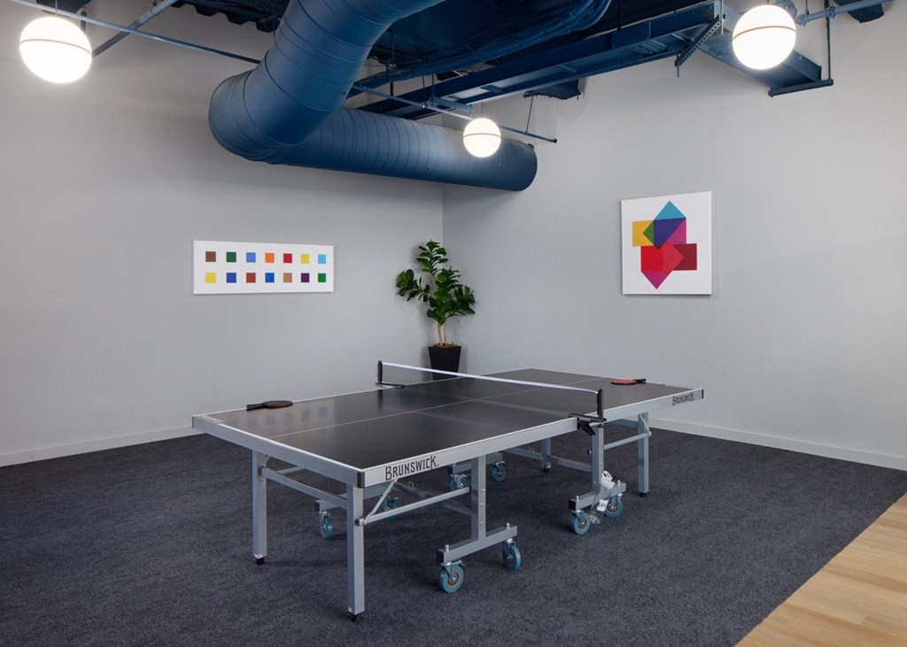 A ping pong table is in the middle of a room with a plant and two paintings on the wall.