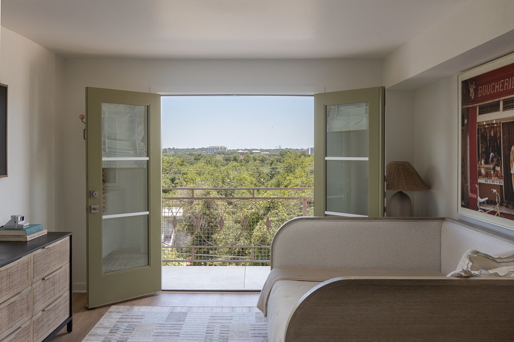 A bedroom with a bed, dresser, and a balcony with a view of the trees at The PARKER on E Mulberry Apartments, San Antonio