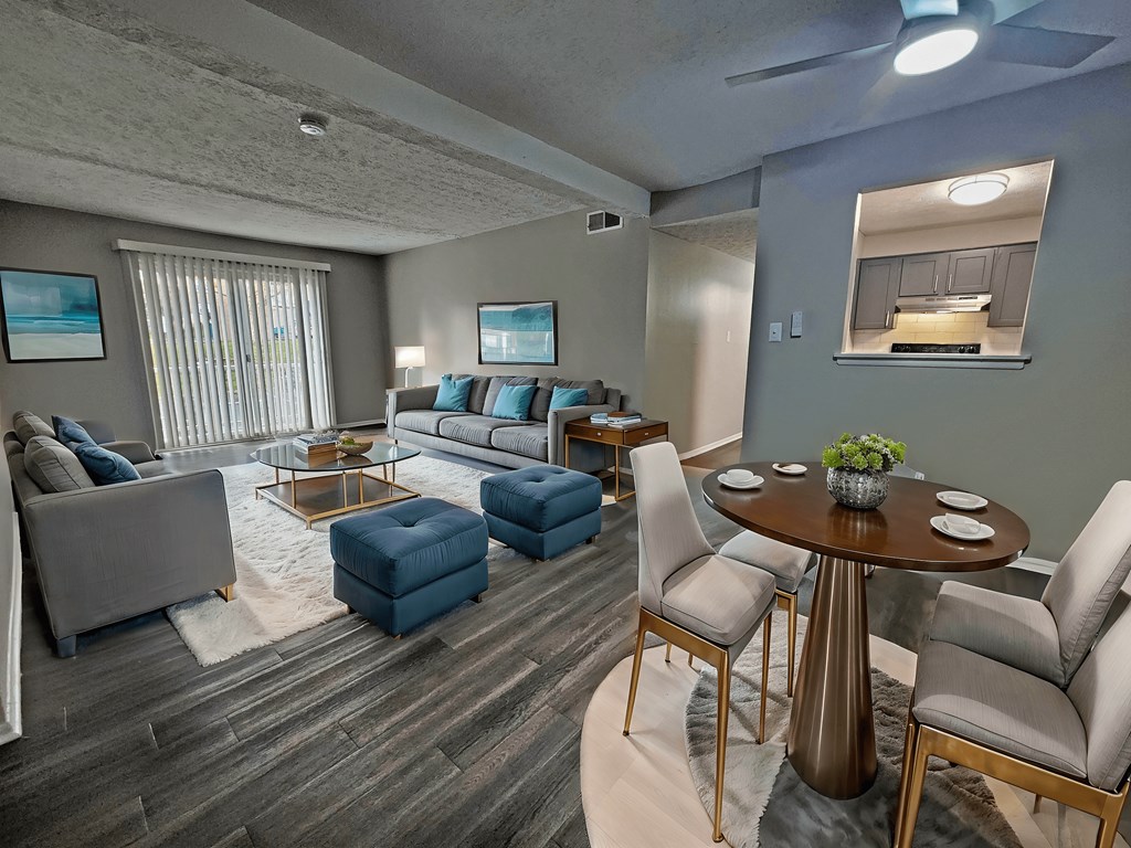 A living room with a grey couch, a blue ottoman, a glass coffee table, and a dining table with chairs.