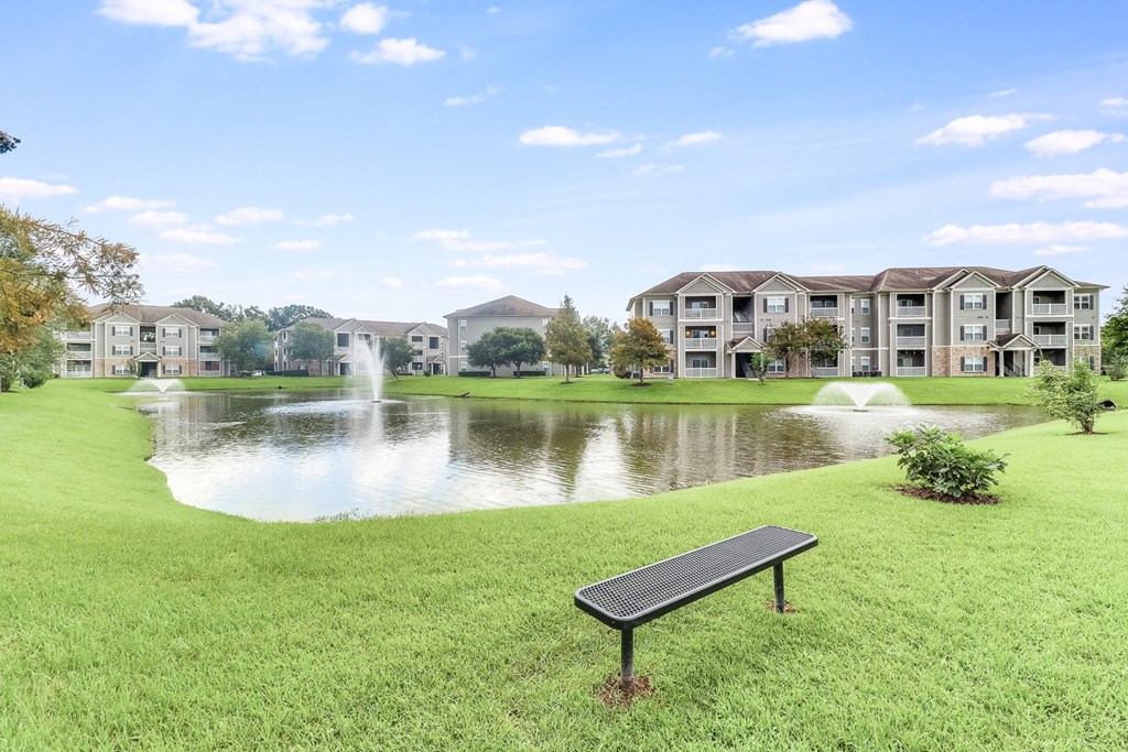 a bench sits in front of a pond with a fountain and apartment buildings in the background at Plantation Crossing, Louisiana, 70508