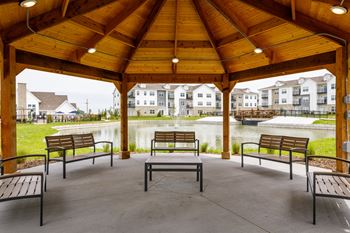 A wooden pavilion with benches is situated in a park with a pond and buildings in the background.