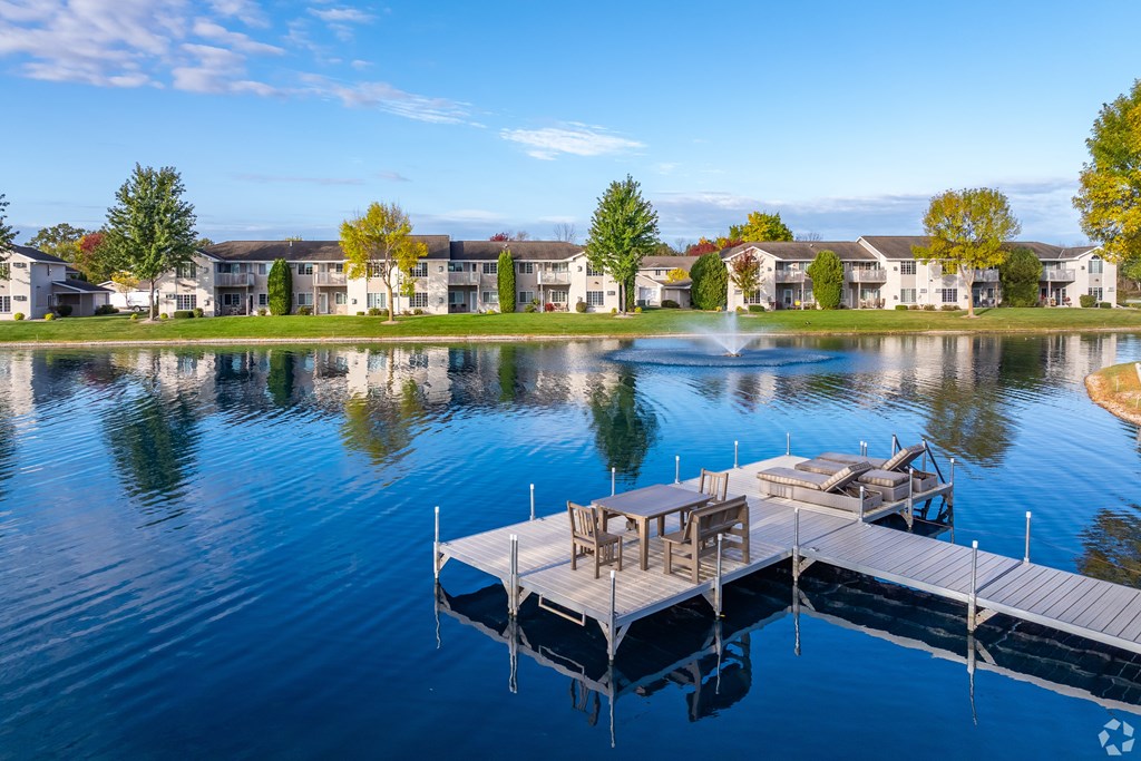 A serene lake with a dock and chairs in the foreground and apartment buildings in the background.